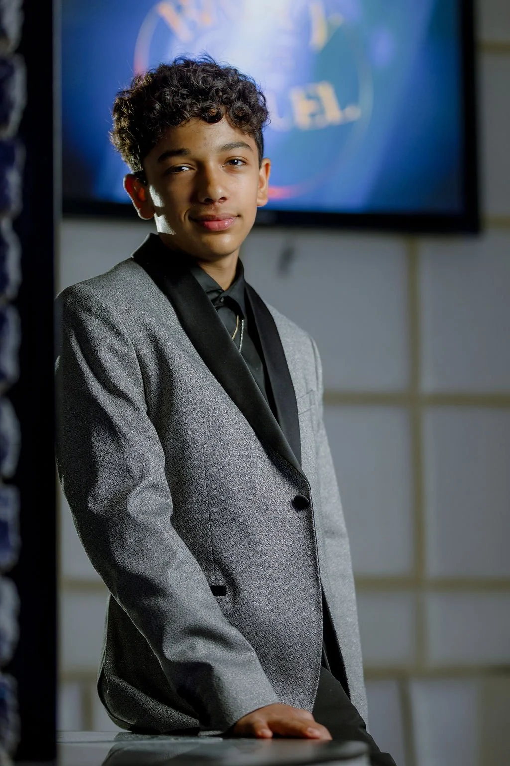 A young man with curly hair wearing a gray blazer and black shirt, standing in front of a television screen with a blurred logo.