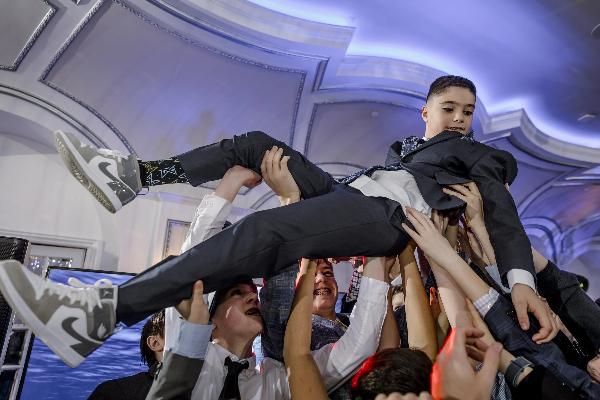 Group of people, including children, lifting a young boy in a suit during a celebration at an indoor event.