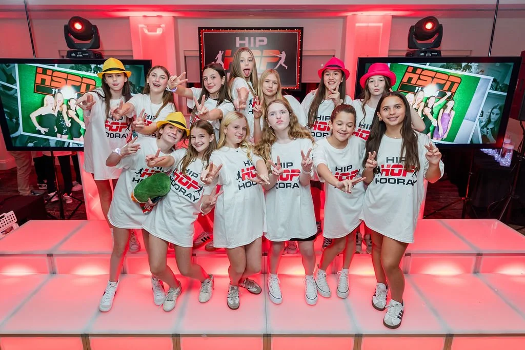 Group of young girls wearing matching white T-shirts with 'HIP HOP' printed on them, posing for a photo on a pink-lit stage with two large screens showing the same logo in the background and two red spotlights overhead.