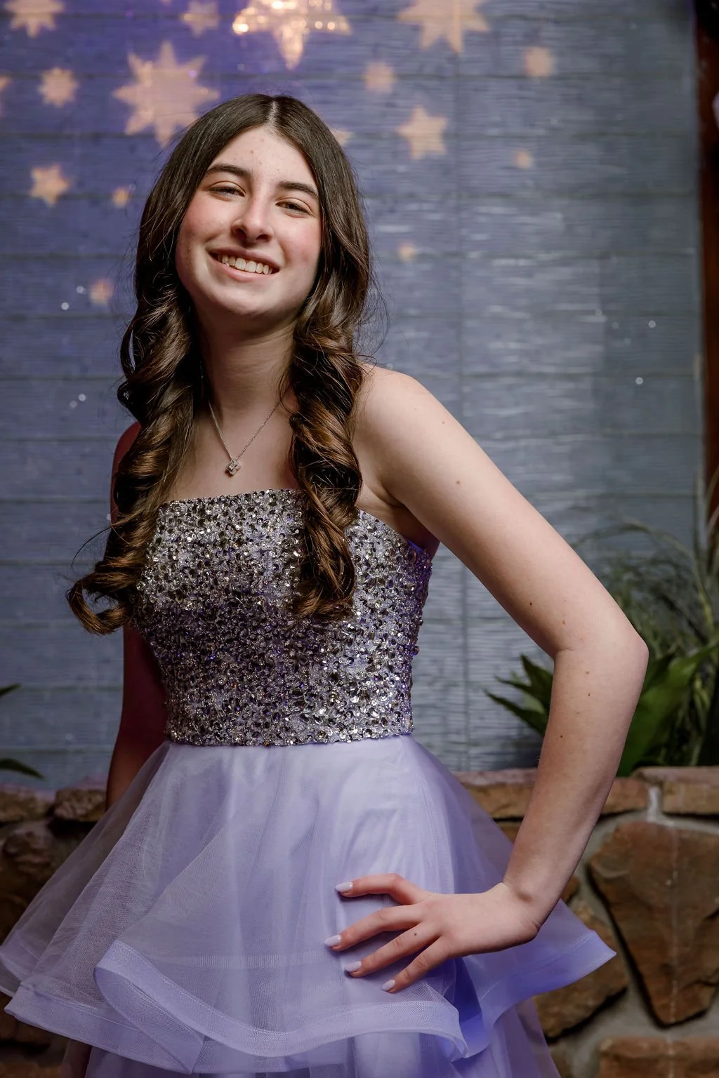 A young woman in a sparkly, strapless dress with a tulle skirt, smiling at the camera with a hand on her hip, standing outdoors against a wooden wall with star decorations.