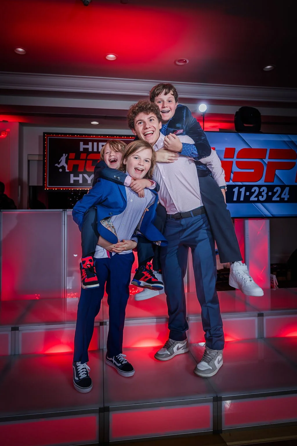 Group of four children, two boys and two girls, smiling and posing joyfully indoors with a sports event banner in the background.