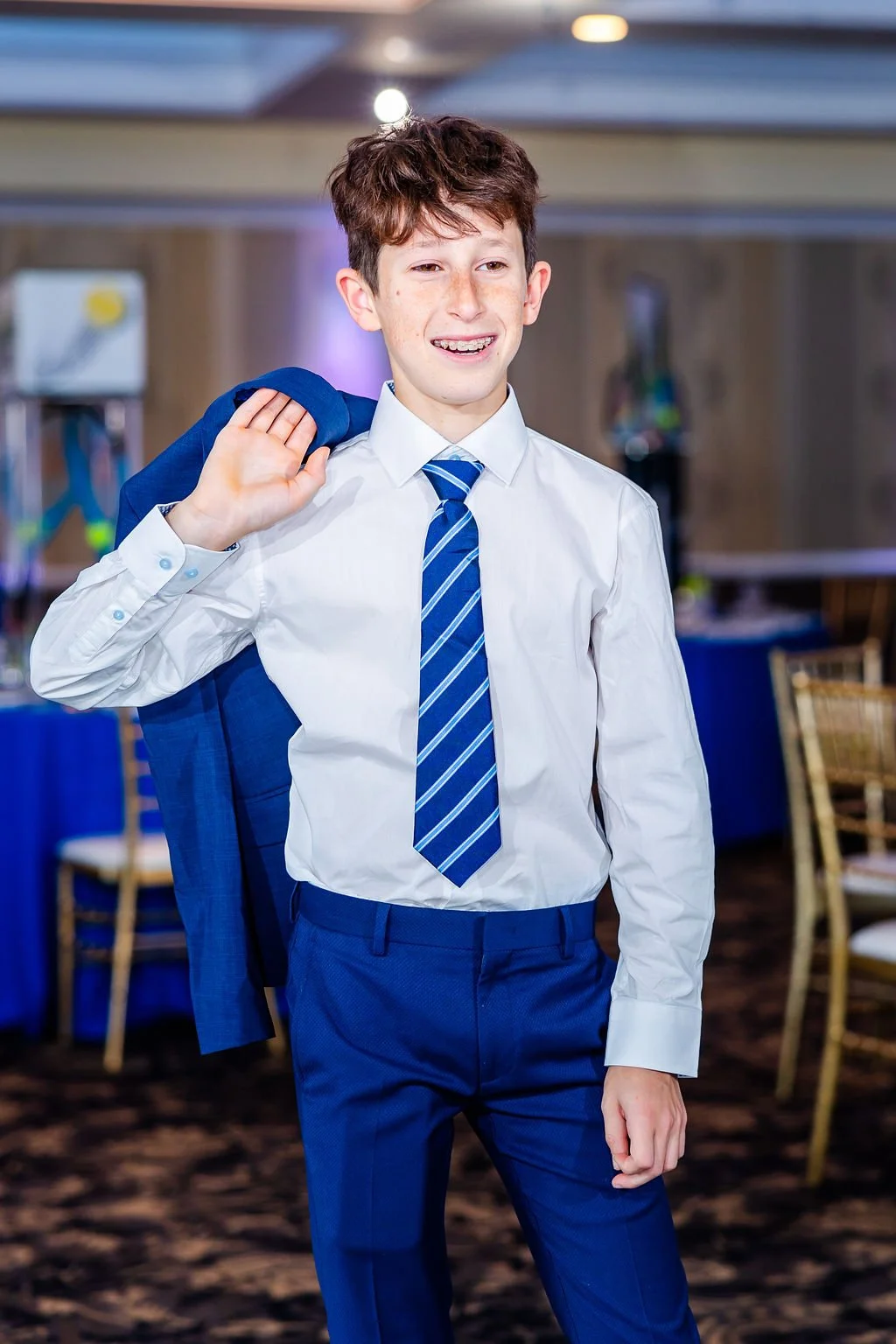 A young boy dressed in a white shirt, blue tie, and blue pants, carrying a blue blazer over his shoulder, smiling in a formal setting.