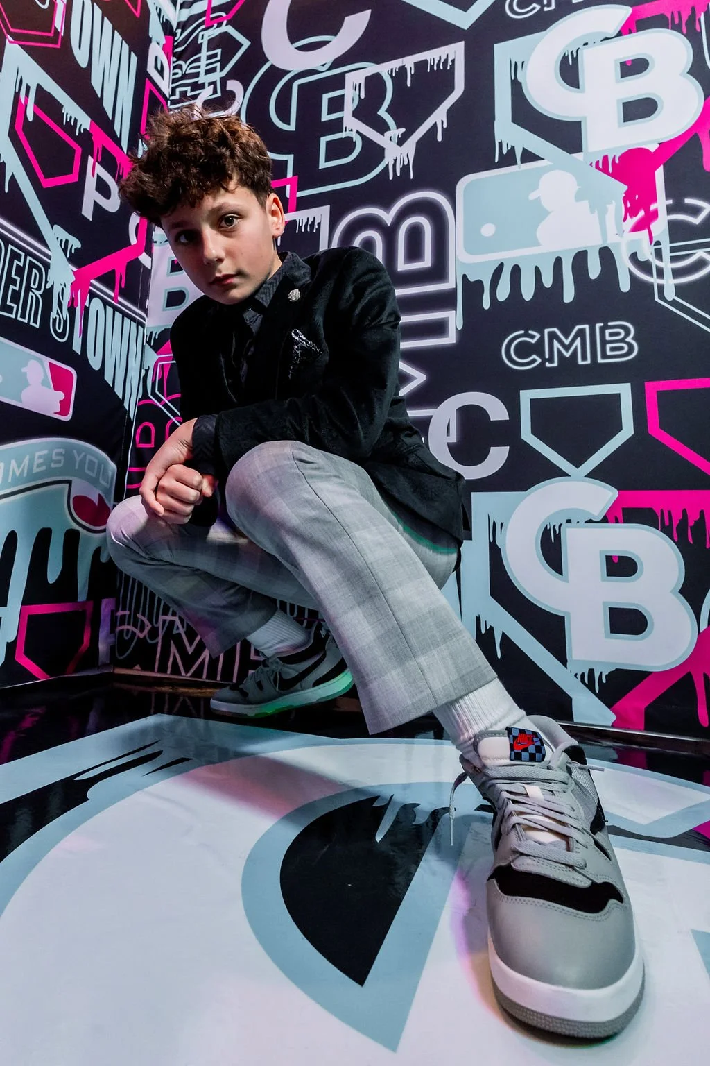 A young boy with curly hair crouching in front of a graffiti-style wall with pink, white, and black graphics including trophies, icons, and text, wearing plaid pants, a black jacket, and Nike sneakers with a Nike logo and checkered design on the tong