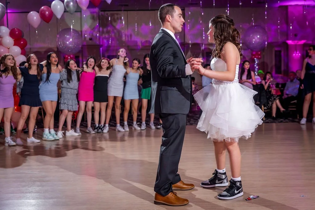 A young girl in a white dress and sneakers is dancing with a man in a suit at a party. Behind them, a group of girls in colorful dresses are watching and smiling. The venue is decorated with pink and white balloons and purple lighting.