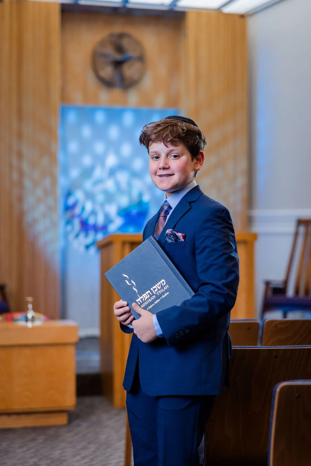 A young boy in a navy suit and tie, holding a program, standing inside a church with wood paneling and chairs in the background.