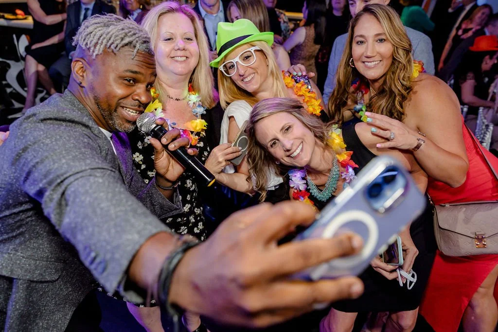 Group of five people taking a selfie at a fun party, wearing colorful leis and accessories, with a crowd in the background.