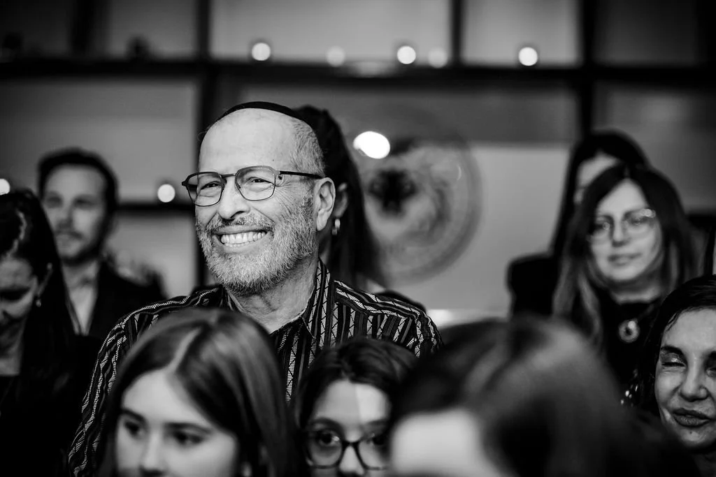 A group of people attending an event, with a middle-aged man with glasses and a beard smiling in the center, surrounded by others in a dimly lit room.