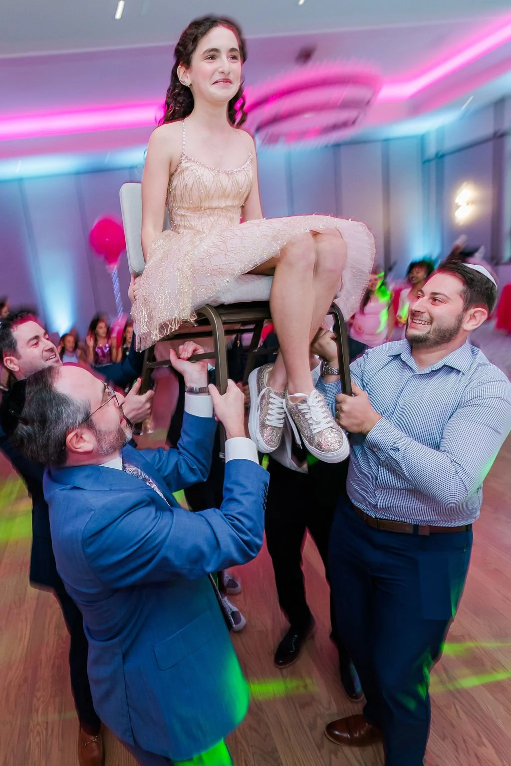 People celebrating at a party, lifting a girl in a dress on a chair in a decorated room with pink and blue lights.