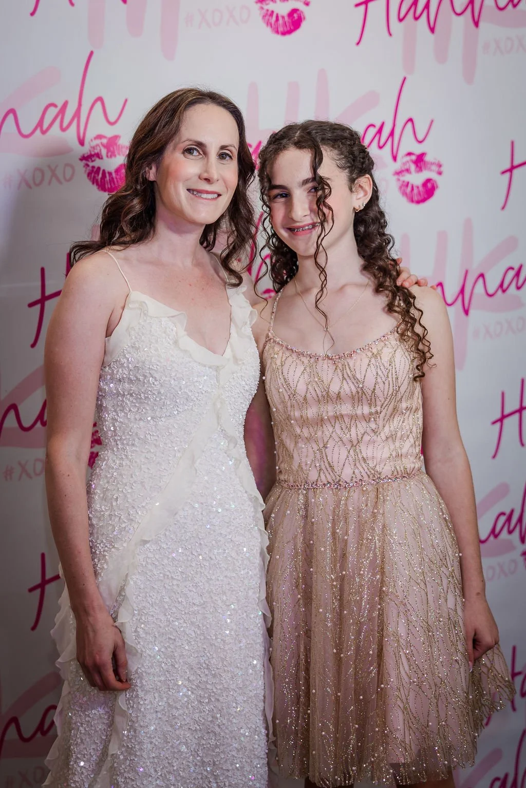 Two women standing close together, smiling for the photo. The backdrop has pink writing and kisses, with the words 'Hannah' and '#XOXO' visible.
