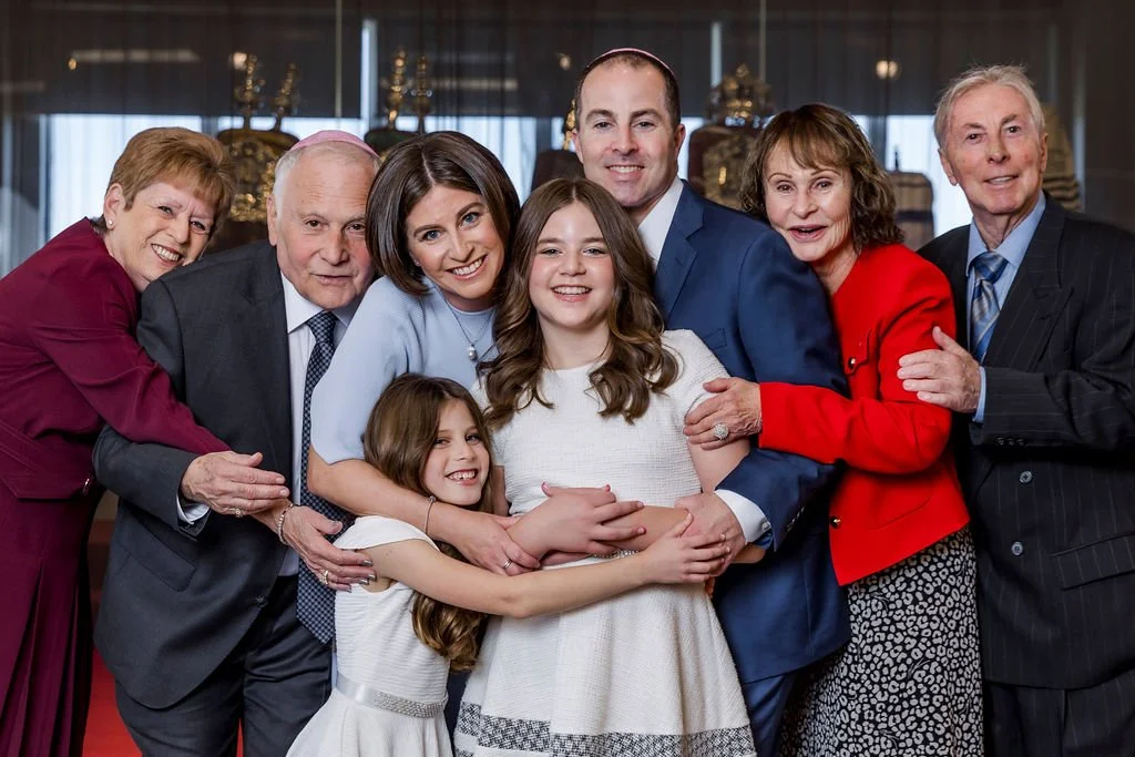A multi-generational family group smiling and hugging at an indoor event, with older and younger adults and children.