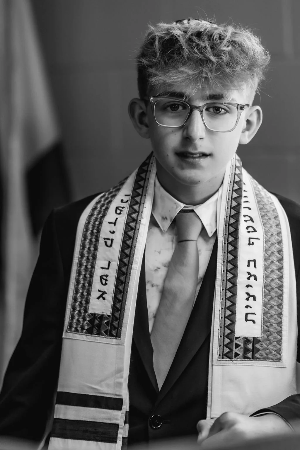 A young man with curly hair and glasses wearing a suit, tie, and a ceremonial sash with Hebrew writing.