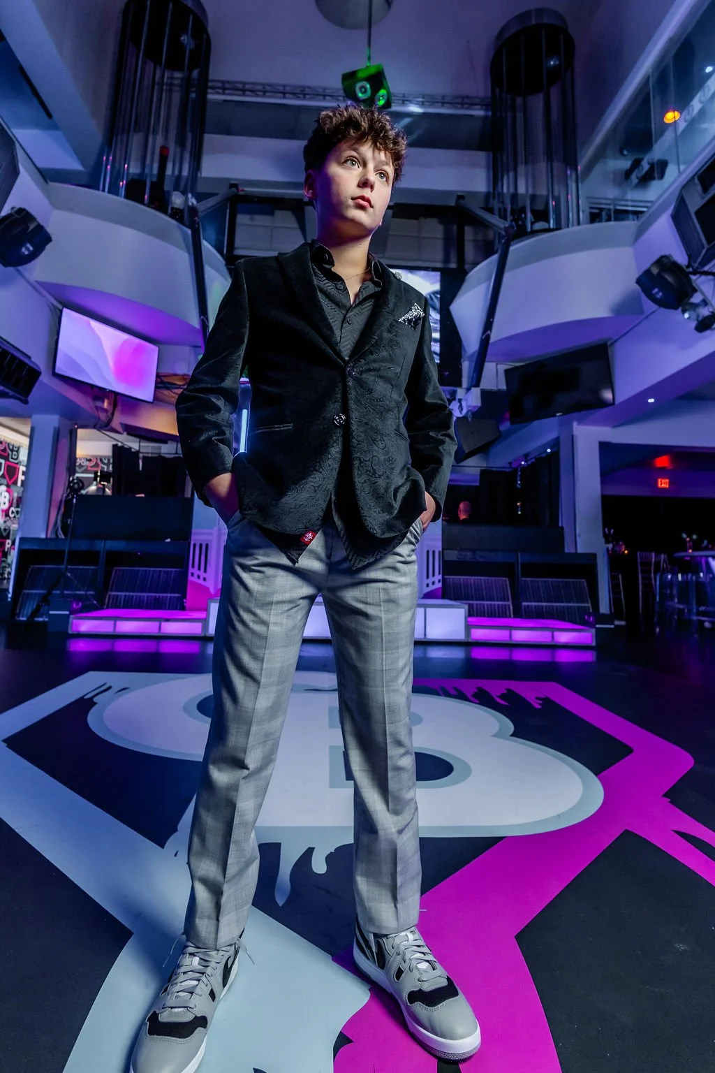 A young person standing confidently in a nightclub with colorful lighting, wearing a black blazer, light-colored checkered pants, and sneakers.