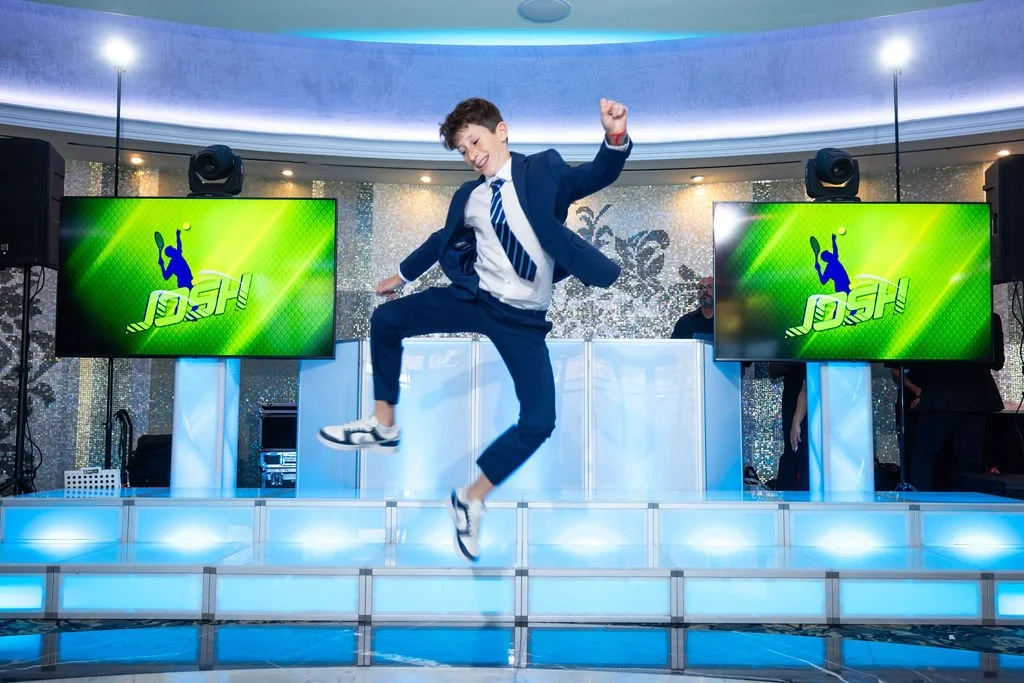 Boy in a suit jumping on a dance floor with DJ setup and two large TV screens in the background displaying a green graphic with the name 'Josh' and a silhouette of a dancer.