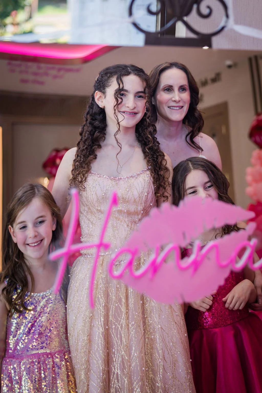A group of five smiling females, including a young girl, two women, and a girl, posing behind a pink decorative sign that says 'Havana'. They are in a decorated indoor setting, dressed in fancy dresses, with balloons and pink decorations around.