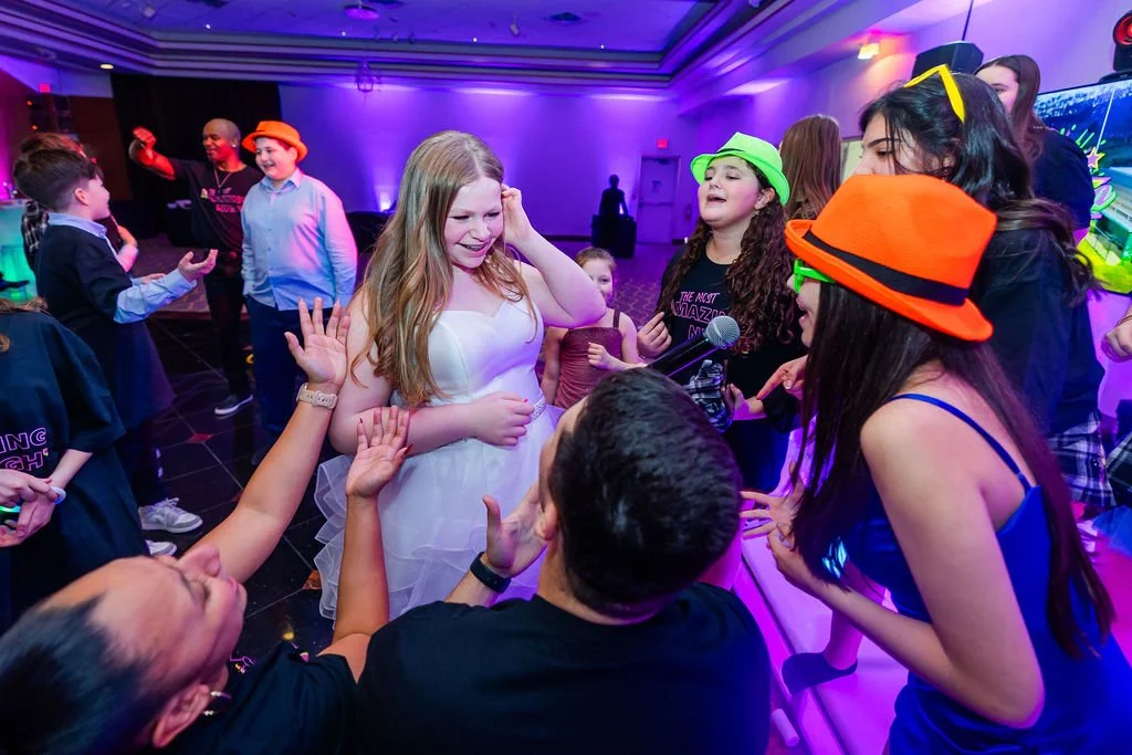 Group of children dancing and singing at a party with colorful hats, a woman with a microphone, and festive lighting.