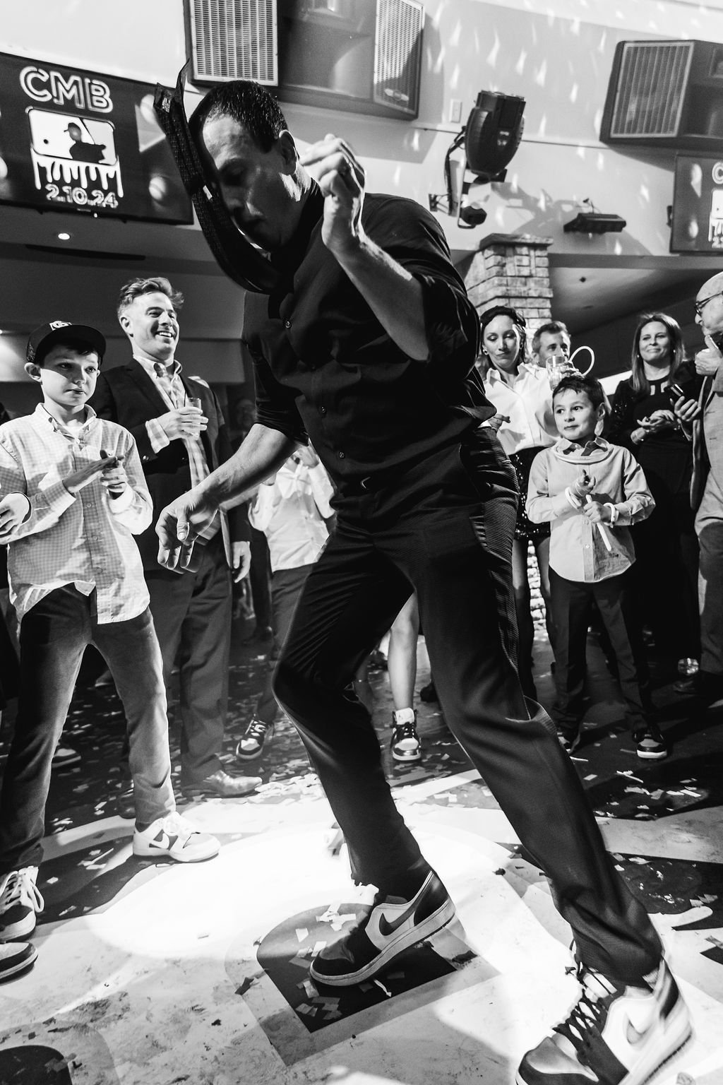 A man dancing on a platform at a party or celebration, surrounded by children and adults watching and smiling.