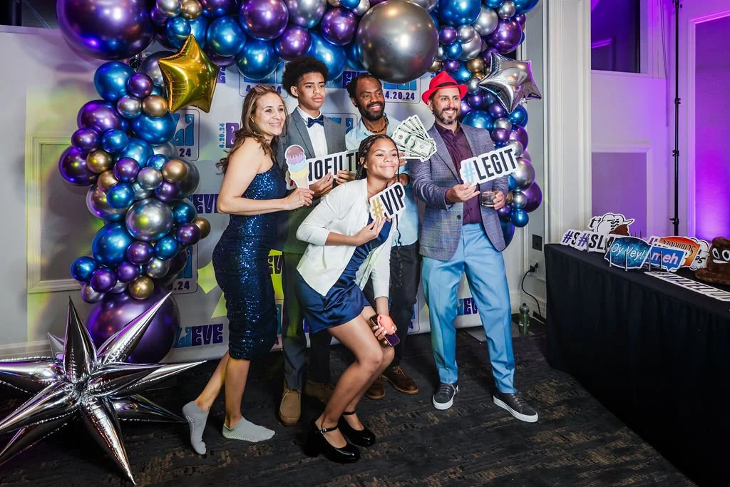 Group of six people dressed in party attire posing at a celebration with purple, gold, and silver balloons, holding signs that read '#VIP,' '#LEGIT,' and '#SLOW,' with a decorated backdrop and table with photo booth props.