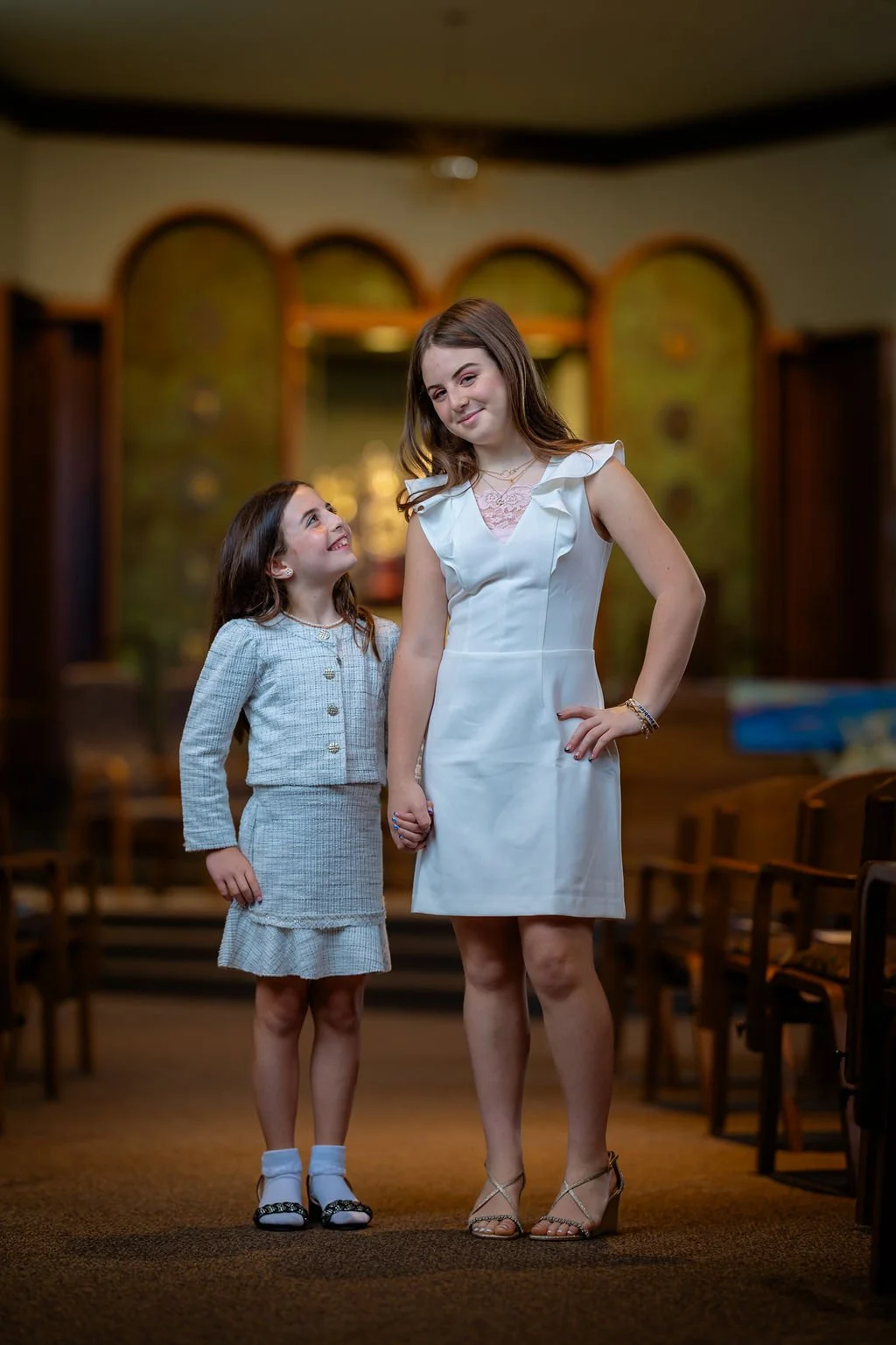 Two young women holding hands, standing in a warmly lit room with wooden chairs, one looking up at the other smiling, the other with a confident smile, dressed nicely.