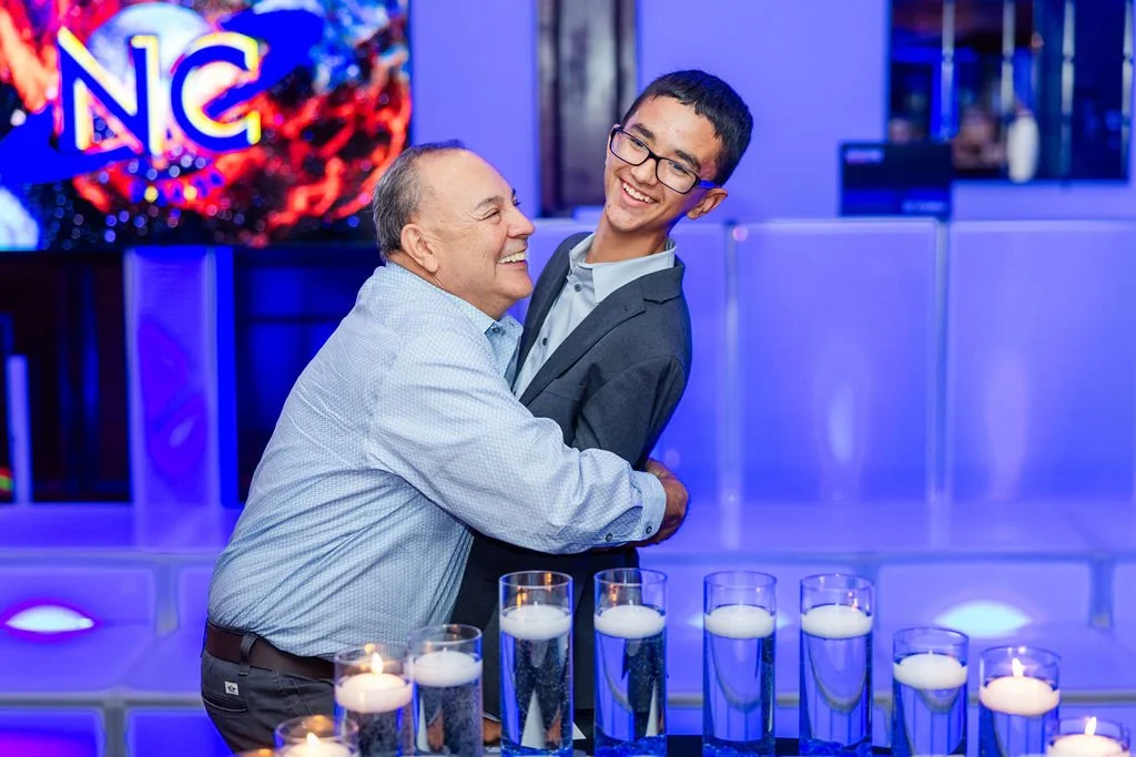 Two men, one older and one younger, are dancing and smiling in a brightly lit, modern reception area with blue lighting. There are multiple glasses of beer on the table in front of them. A large colorful 'NC' logo is visible in the background.