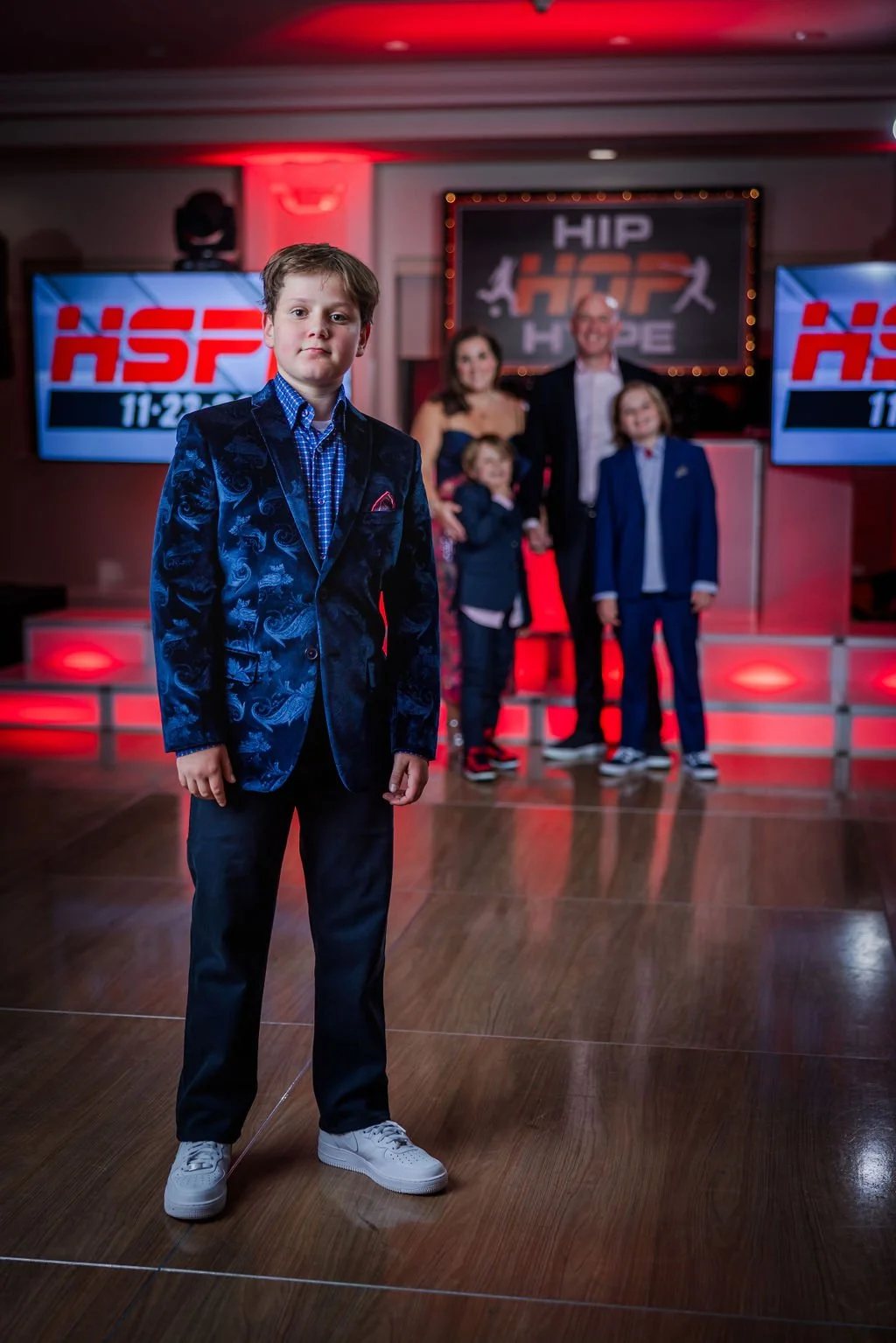 A young boy in a dark blue patterned blazer and white sneakers standing in front of a family at an event, with two digital screens displaying the letters "HSF" and a large screen with the logo "HIP HOP HOPE" in the background.