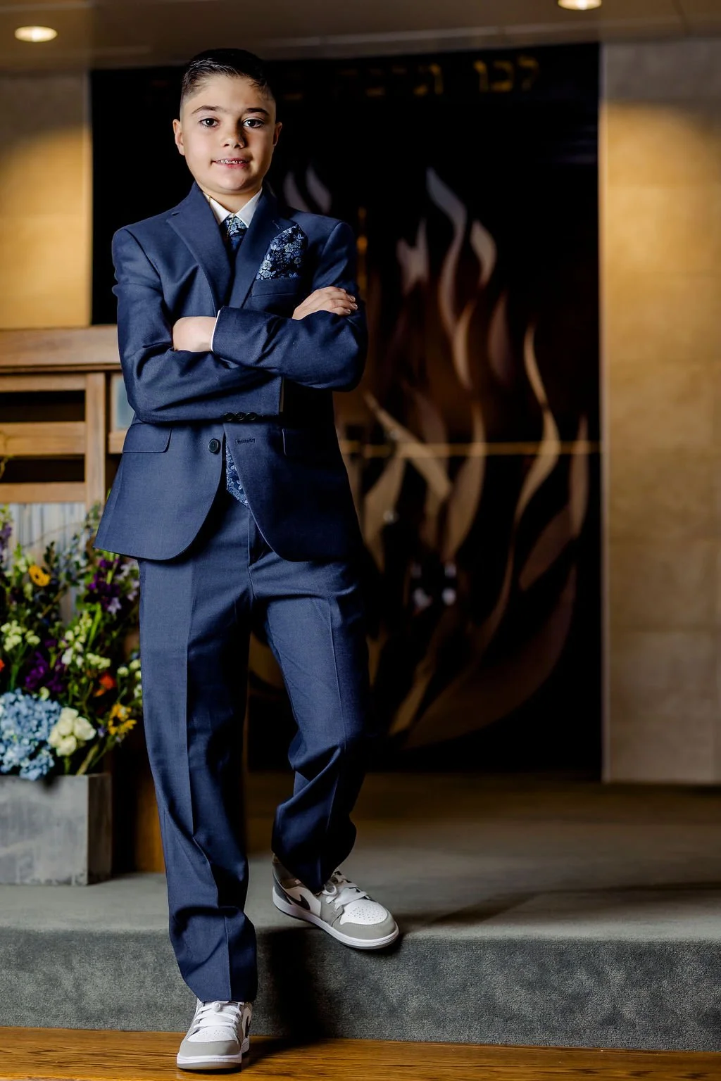 Young boy wearing a dark blue suit with a tie, standing with arms crossed in an indoor setting with a dark background and floral arrangement nearby.