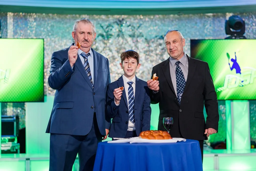 Three men in suits and a boy in a suit standing behind a table with a loaf of bread, holding slices of bread. The background shows bright lights, two green screens with tennis-themed graphics, and sparkling decorations. All are posing for a photo, po
