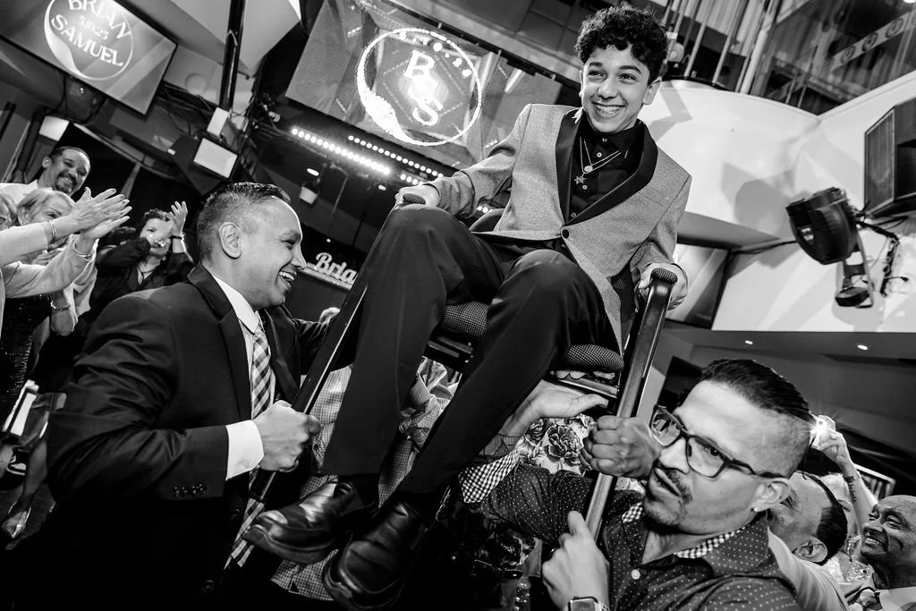 A young man in a suit sitting in a chair being lifted by two men at a celebration or party, with other happy people clapping and smiling in the background.