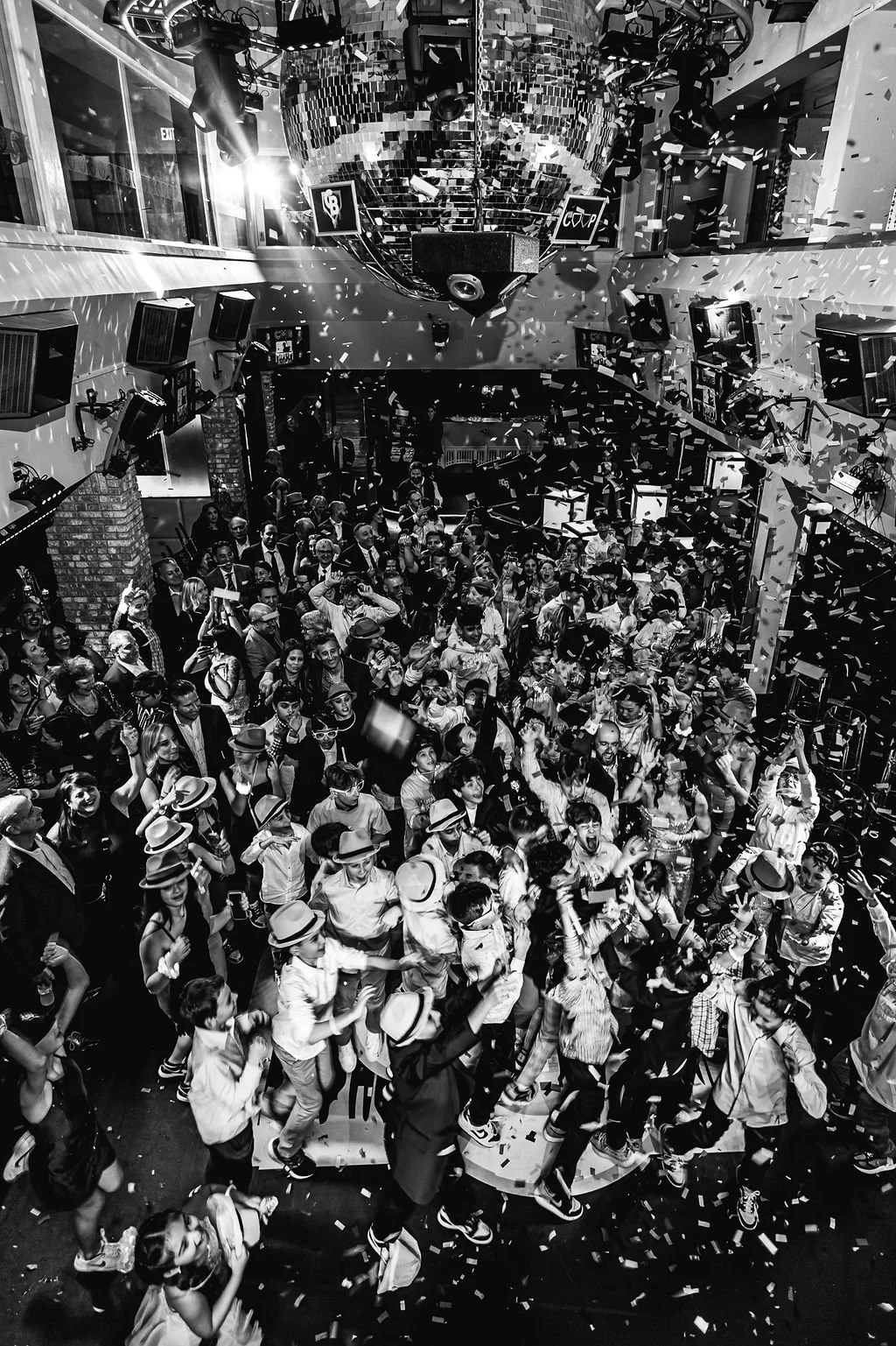 A large crowd of people dancing and celebrating at an indoor party with a disco ball, confetti falling, and stage lighting.