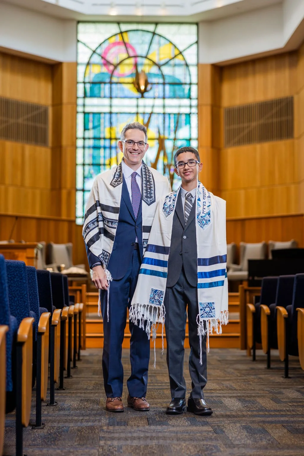 Two men standing in a synagogue, wearing traditional Jewish prayer shawls called tallit, smiling at the camera with a stained glass window in the background.
