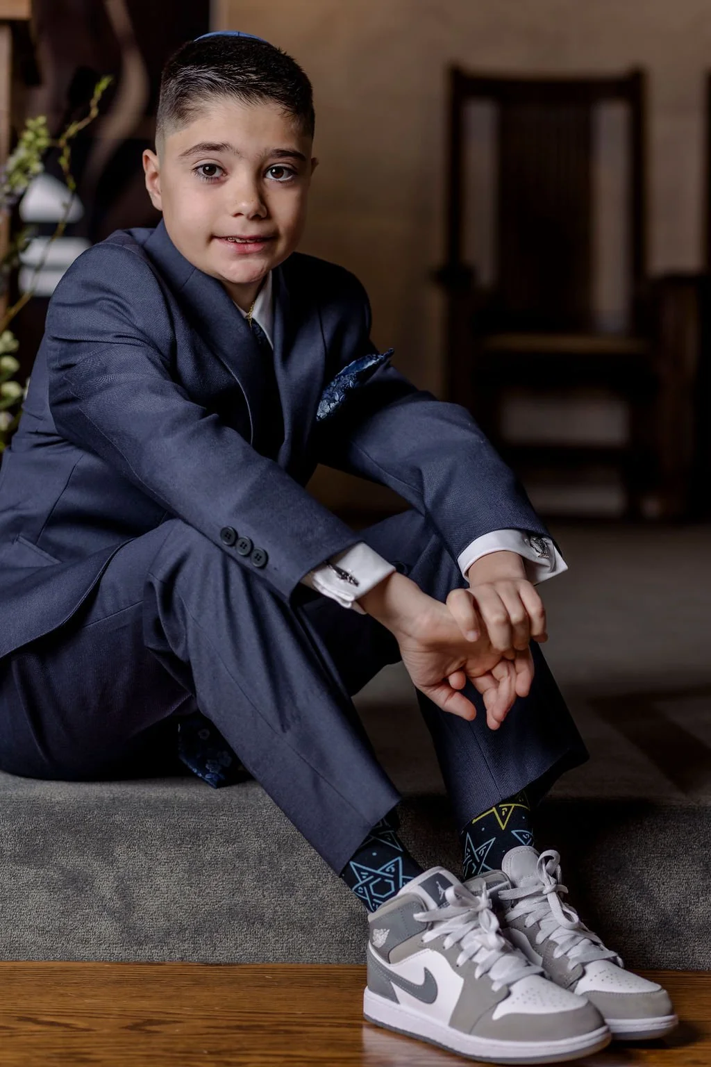 A young boy dressed in a navy suit with white shirt, wearing patterned black and blue socks, and gray and white sneakers, sitting on a low surface in an indoor setting.