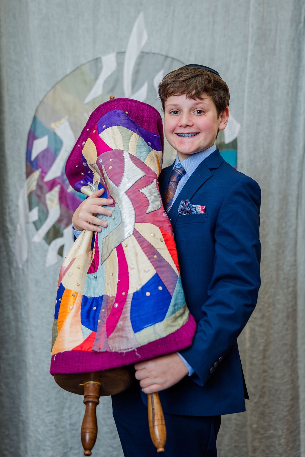 A young boy dressed in a blue suit, smiling and holding a colorful quilted object with wooden legs.
