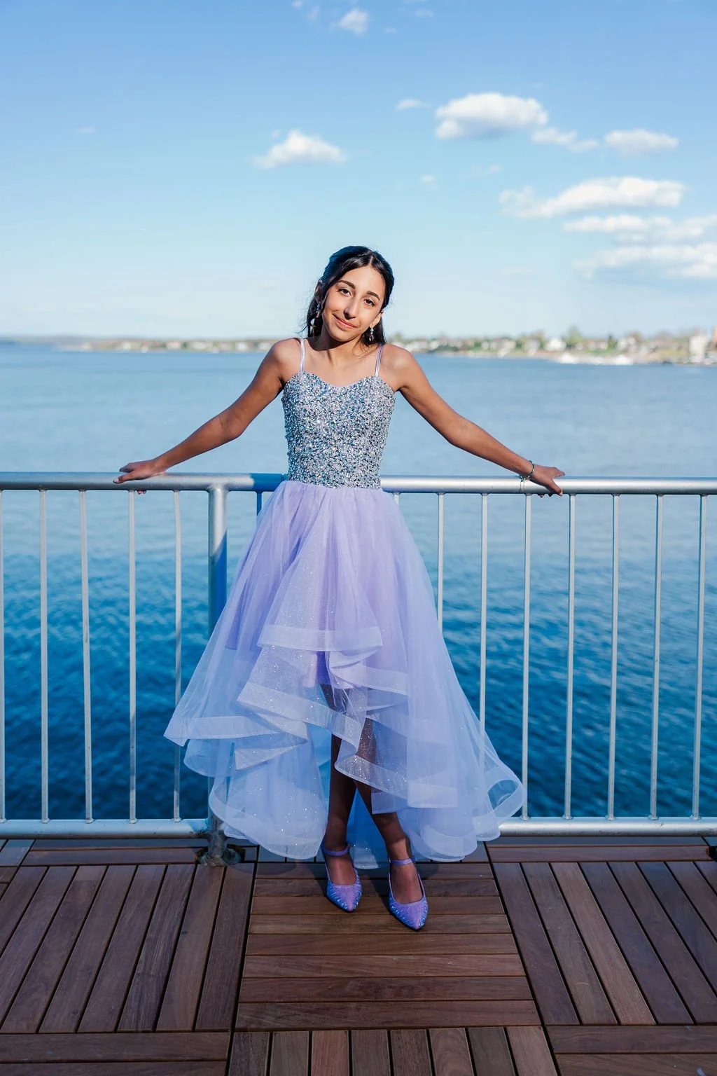 A young woman in a sparkly dress standing on a boat deck by the water, smiling at the camera with arms outstretched on a metal railing, with a city skyline and blue sky with clouds in the background.