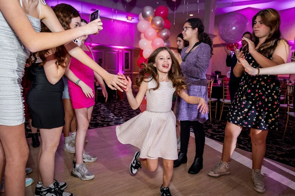 A girl is dancing joyfully at a party surrounded by other girls, some taking photos and others clapping, with pink and purple balloon decorations in the background.