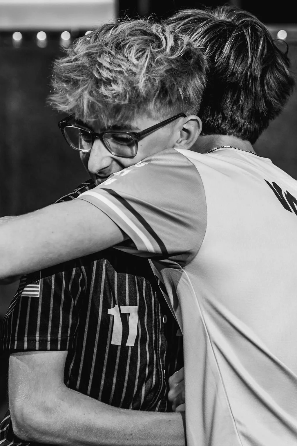Two young men hugging, one wearing glasses and a striped jersey, the other in a plain sports shirt, in a black-and-white photo.