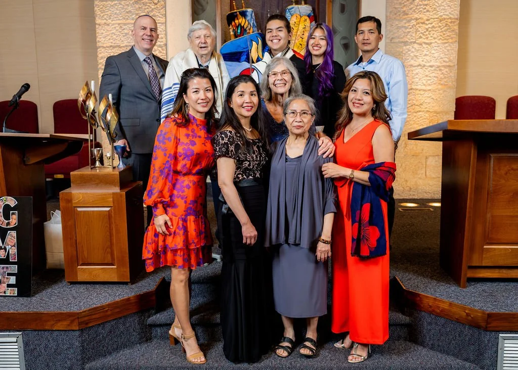 A group of eleven people, men and women, standing together in a formal setting, smiling at the camera. They are dressed in colorful and formal attire, with a backdrop of a stage and flags or banners.