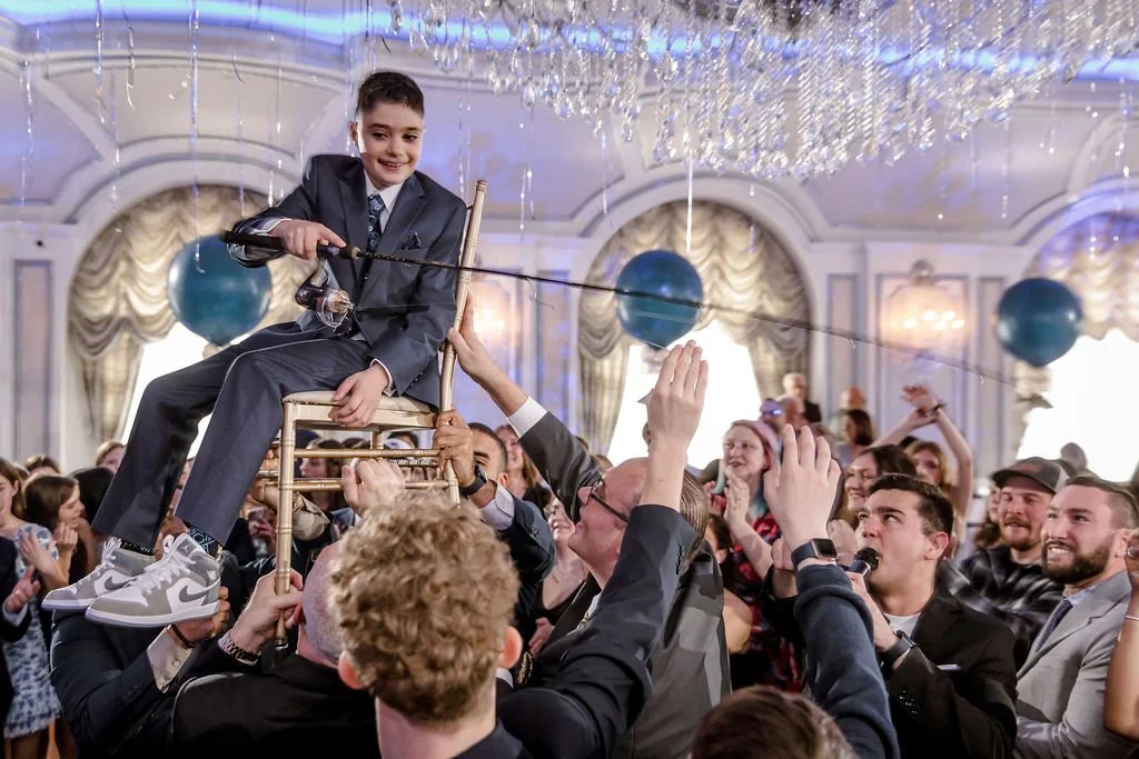 A young boy in a suit sitting on a chair held up by adults, in a festive indoor setting with a chandelier, decorations, and a crowd cheering.