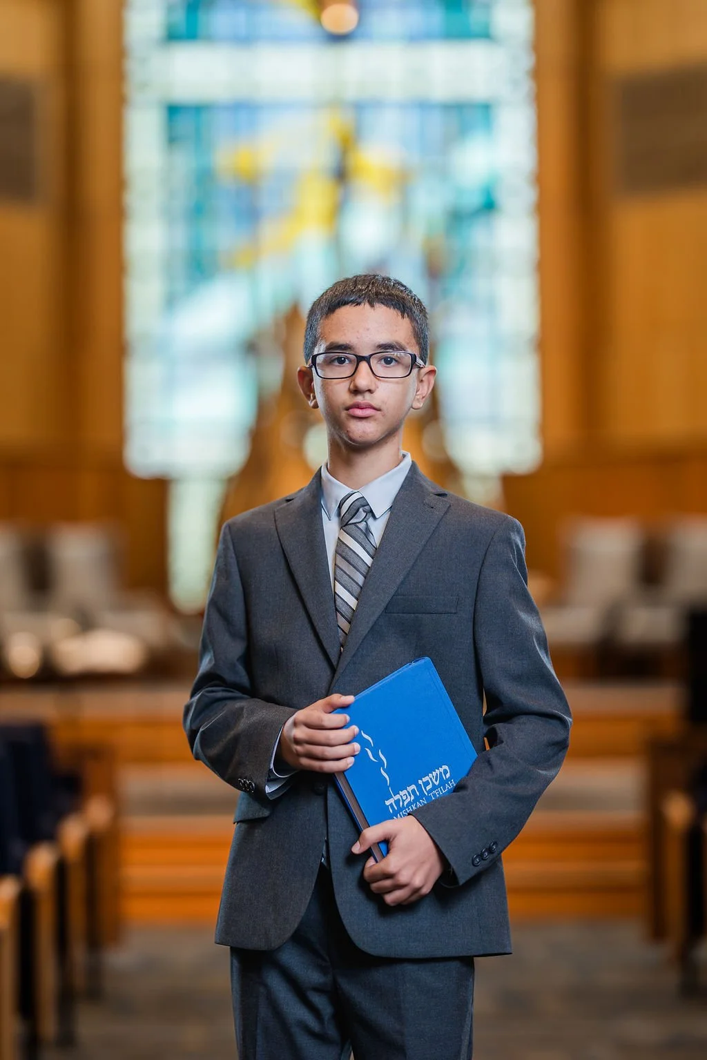 Young boy in a suit holding a blue book inside a church, with stained glass windows in the background.