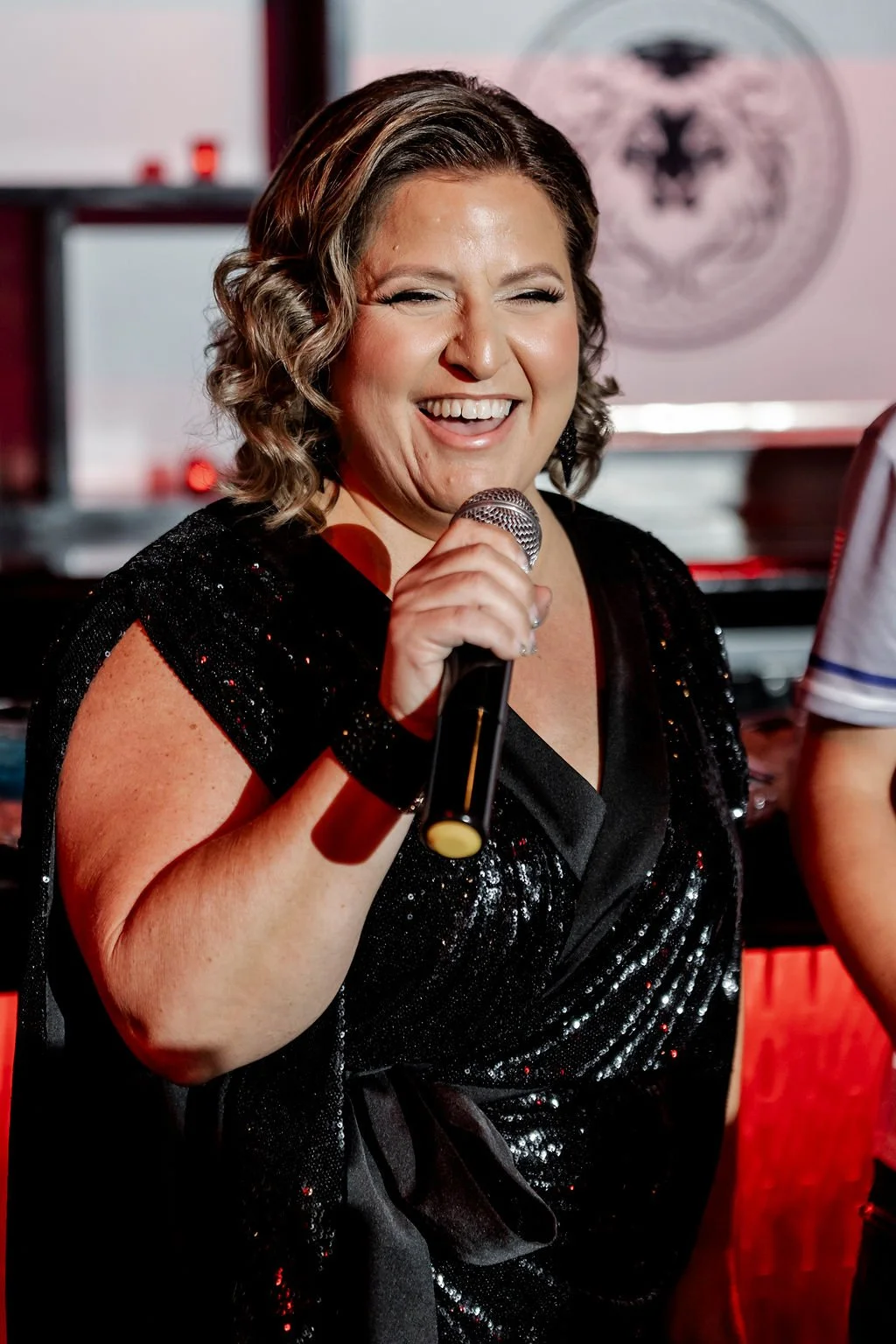 A woman with short wavy hair, wearing a black sequined dress, smiling, and holding a microphone, possibly singing or speaking.