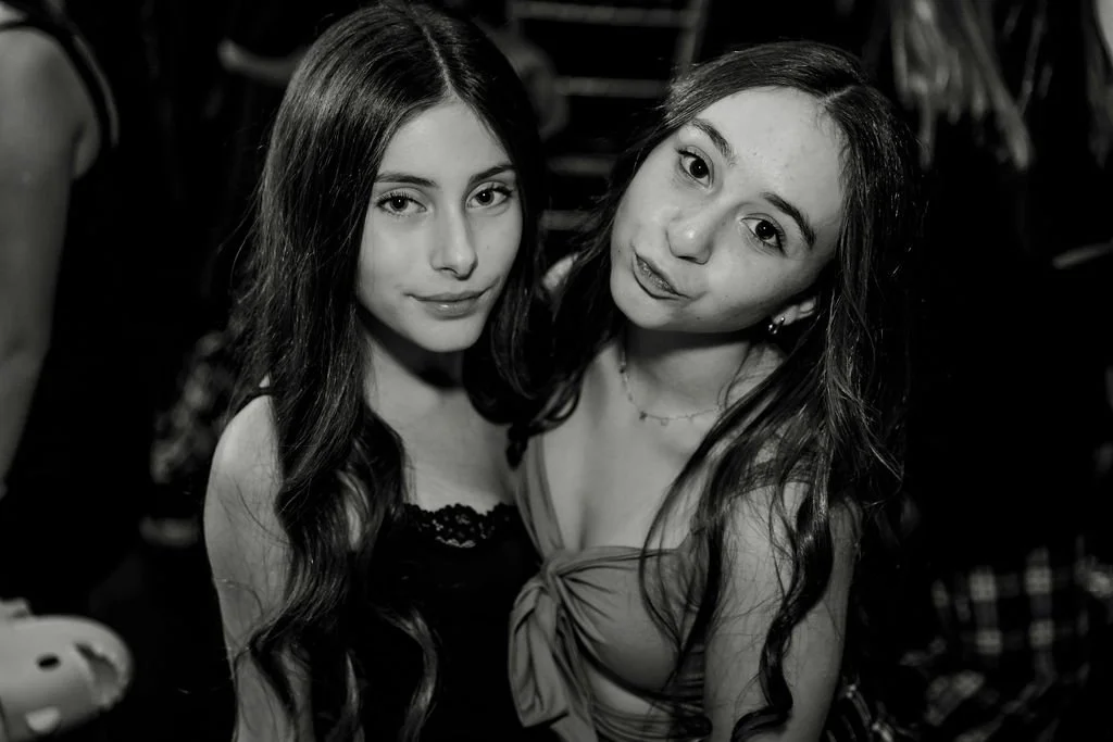 Two young women with long hair posing for a photo in a black-and-white setting. One is wearing a black top with lace details, and the other is wearing a dress with a bow on the front.