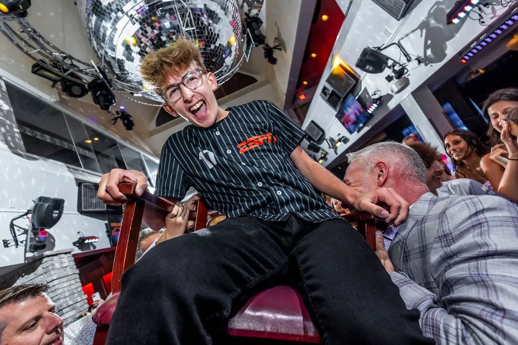A young man with glasses laughing on a chair being lifted at a lively party or event, surrounded by smiling people in a decorated indoor venue, with disco balls and lighting.