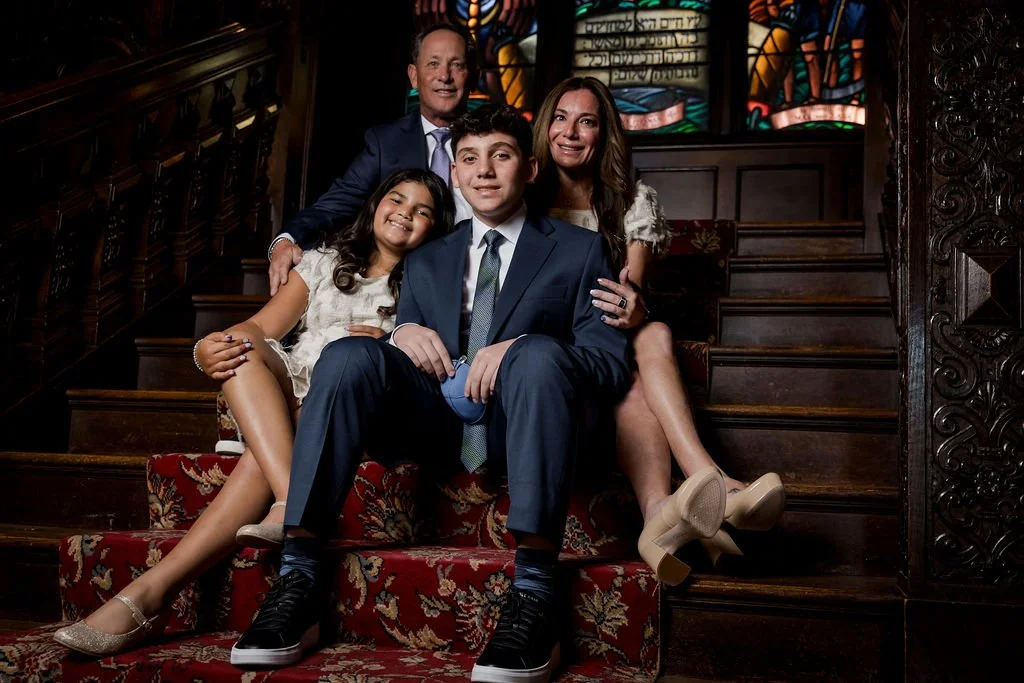 A family of four sitting on a staircase in a church, with stained glass windows behind them. The parents are seated at the top, with the mother wearing a white dress and beige heels, and the father in a dark suit. Their children, a teenage boy in a s
