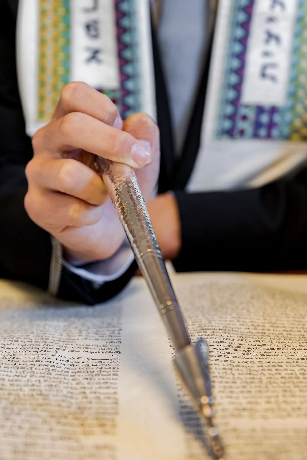 Close-up of a person holding a silver pen and pointing at a Hebrew text in a book.