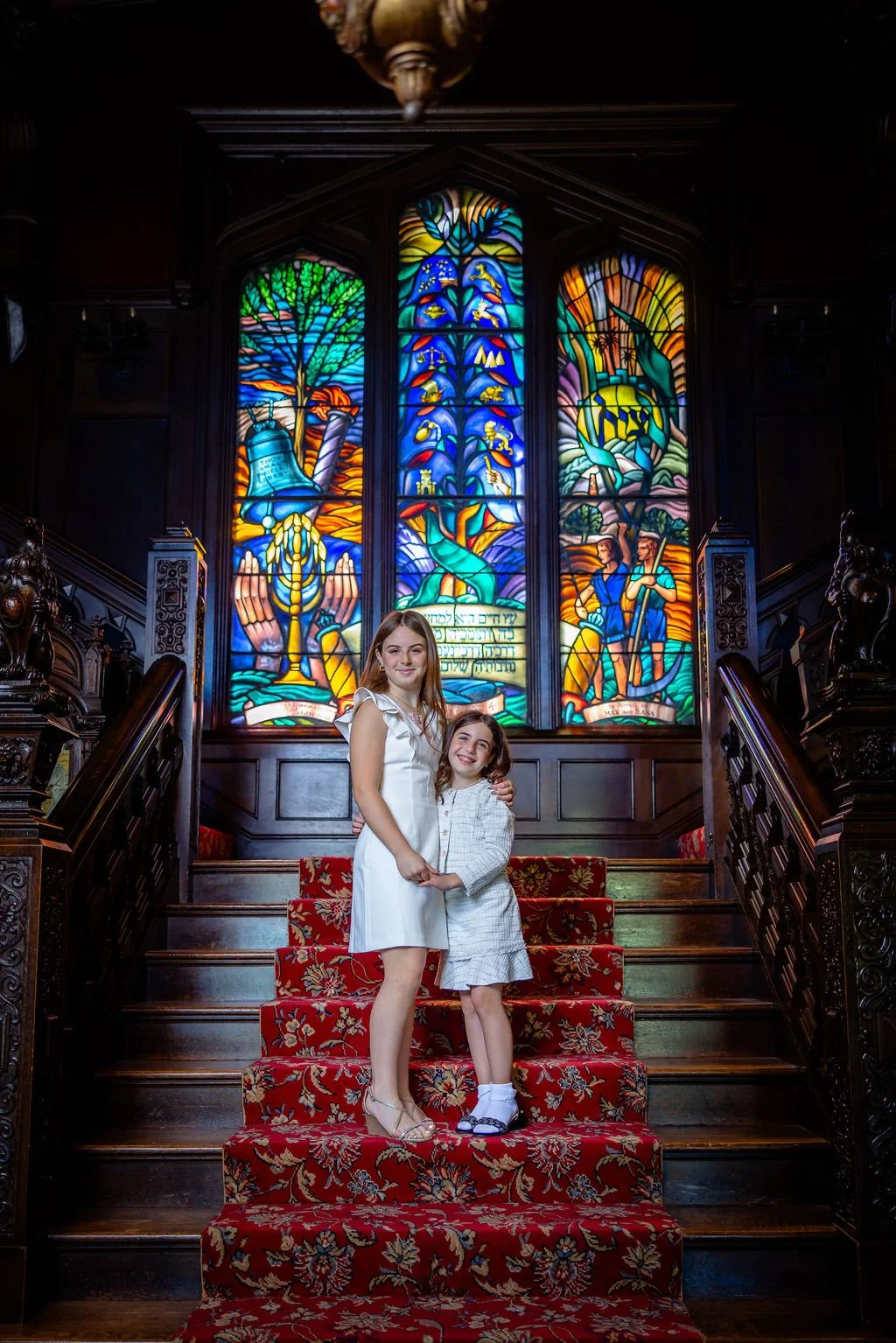 Two smiling girls, one older and one younger, standing on a red-carpeted staircase in front of a large colorful stained glass window depicting biblical or religious scenes, inside a church or chapel.