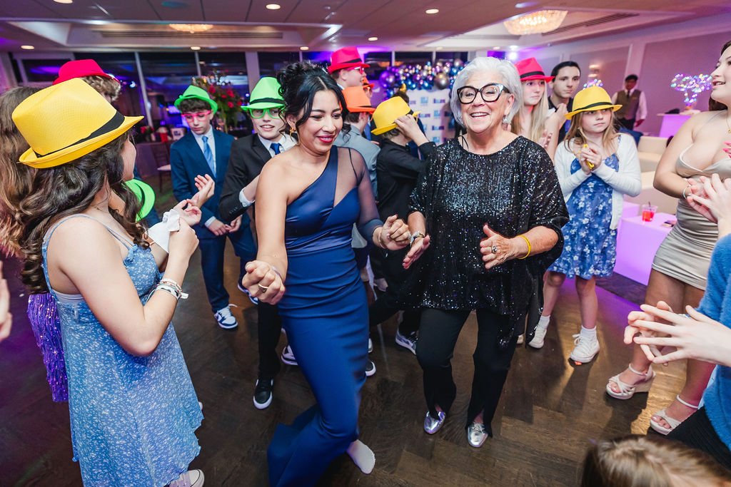 People dancing at a celebration party with colorful hats and party outfits, in a decorated indoor venue with purple lighting.