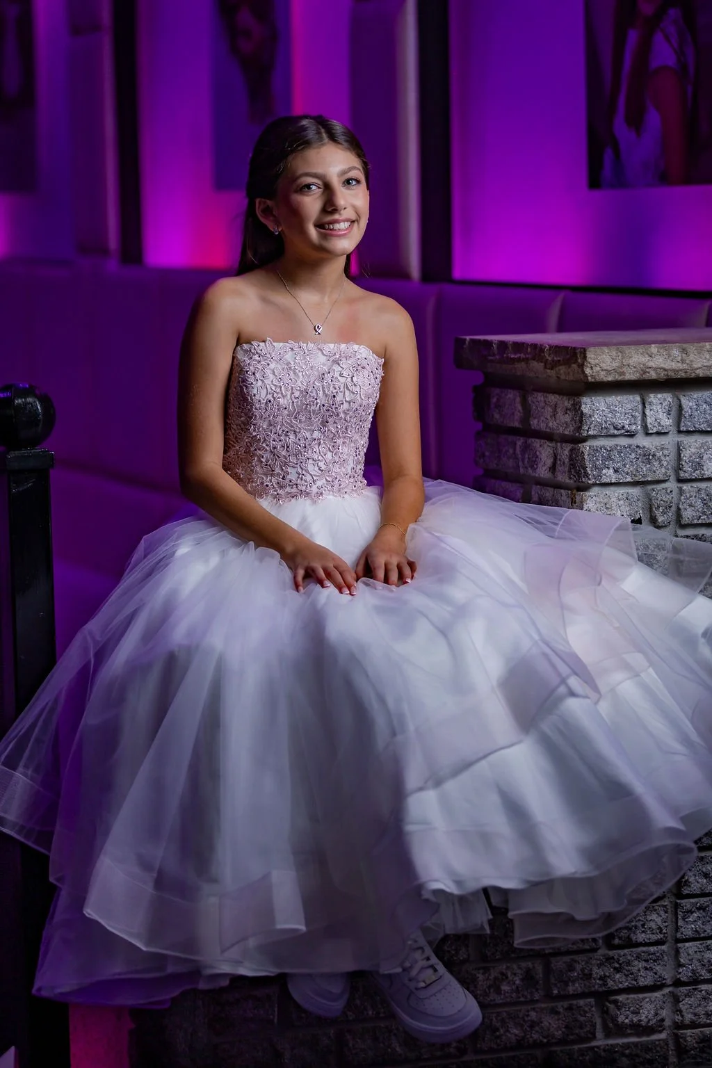 Young girl in a pink lace top and white tulle dress sitting on a ledge in a dimly lit venue with purple lighting.