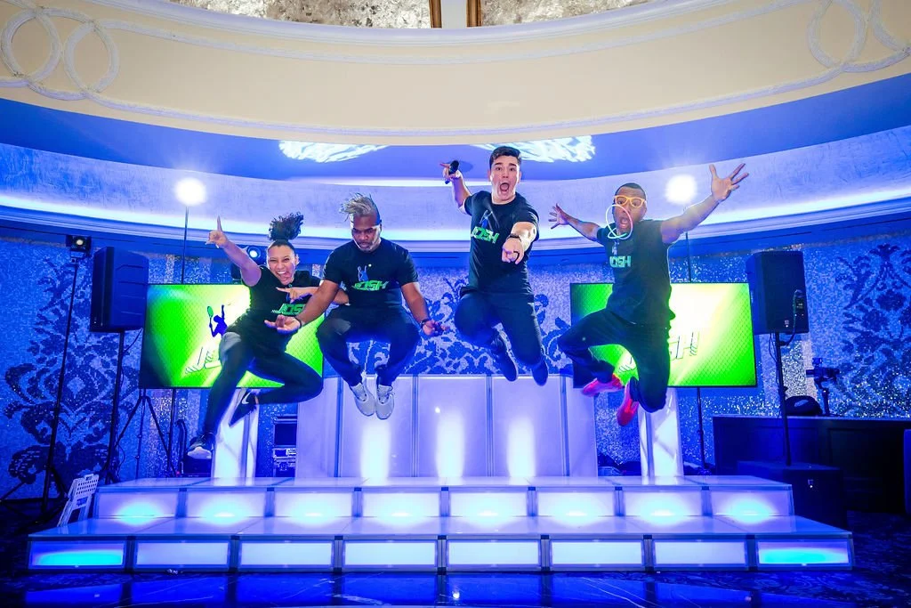 Four people jumping in the air on a stage with colorful lighting, large screens, and patterned walls in the background.