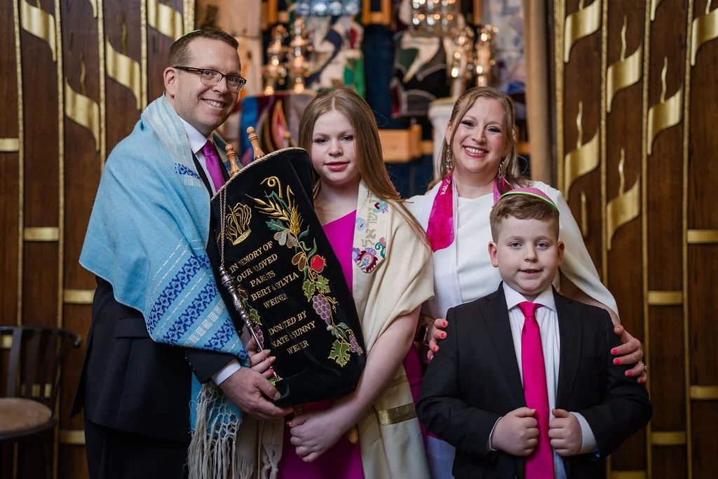 A family celebrating a Jewish religious ceremony, with a man and a woman, two children, and a Torah scroll, inside a synagogue with wooden decor.