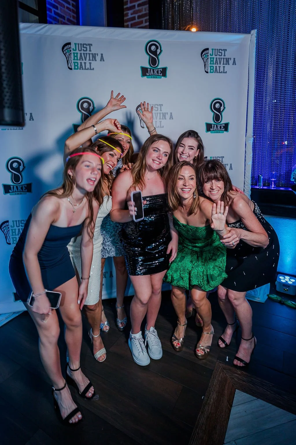 Group of women celebrating at a party, posing in front of a photo backdrop with logos and text, some wearing colorful headbands and holding smartphones.