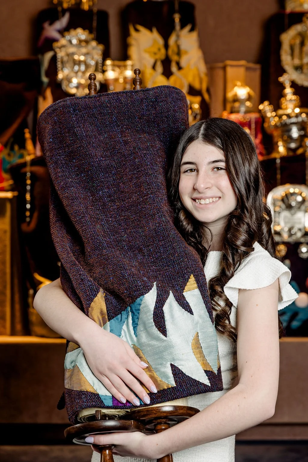 A young girl with long, dark curly hair is smiling and hugging a large, colorful woven quilt with a sailboat design. She is indoors with shelves of decorative items and clocks in the background.