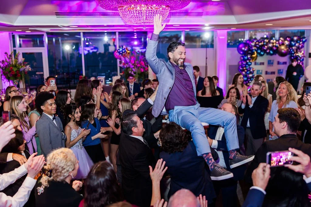 A man is lifted on a chair during a celebration or dance at a decorated event, surrounded by smiling guests, colorful balloons, and floral arrangements.
